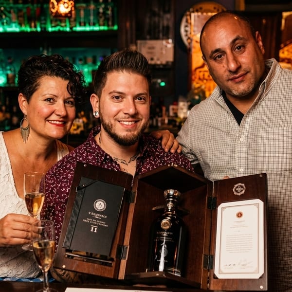Three smiling people pose with a bottle of V.S. Silevracso II in a wooden box at a bar.