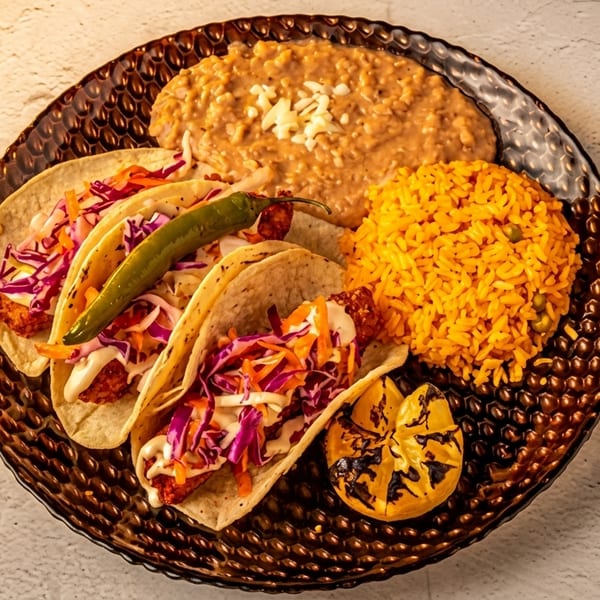 Three fish tacos with cabbage slaw, a green chili, rice, beans, and grilled lemon on a dark plate.