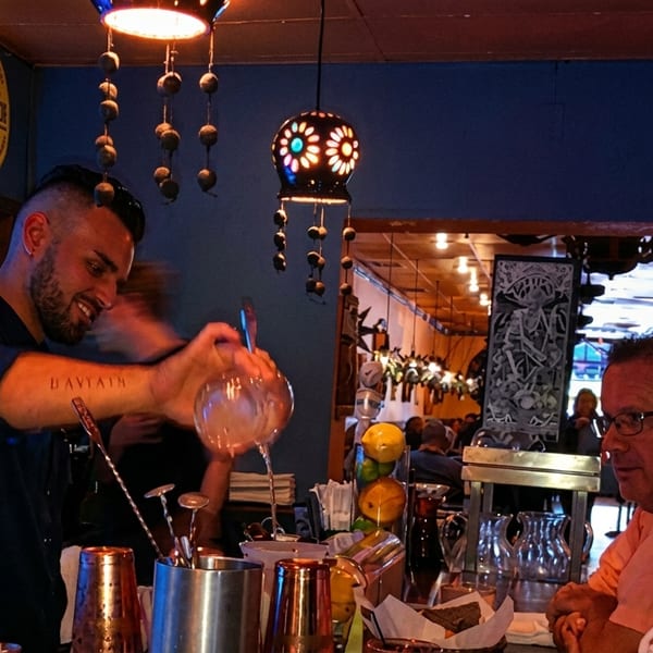 A bartender mixes drinks for two customers at a lively, dimly lit bar.