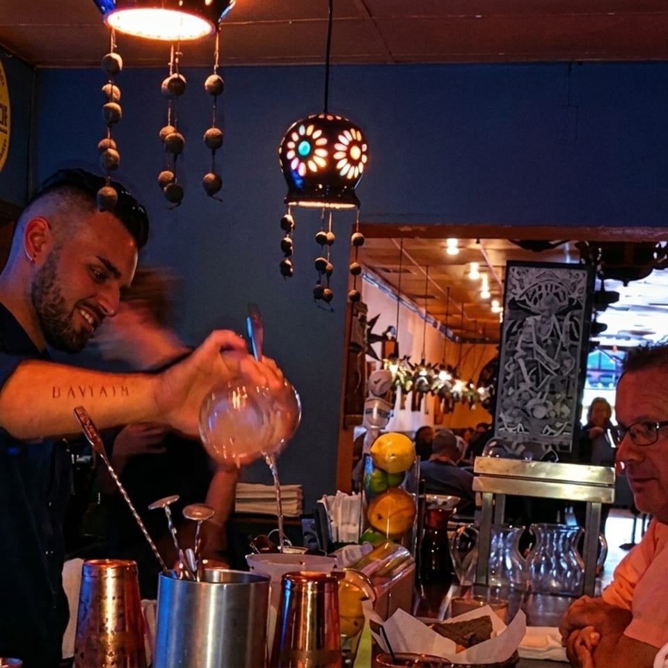 A bartender mixes drinks for two customers at a lively, dimly lit bar.