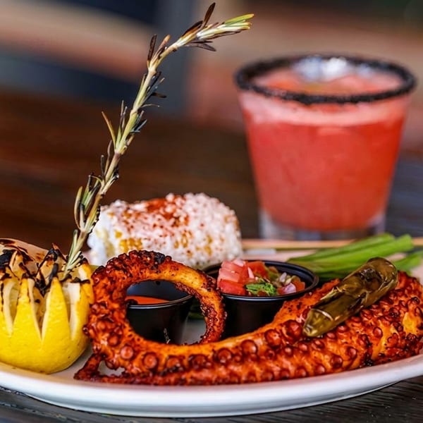 Grilled octopus with corn, lemon, sauces, and a red cocktail on a wooden table.