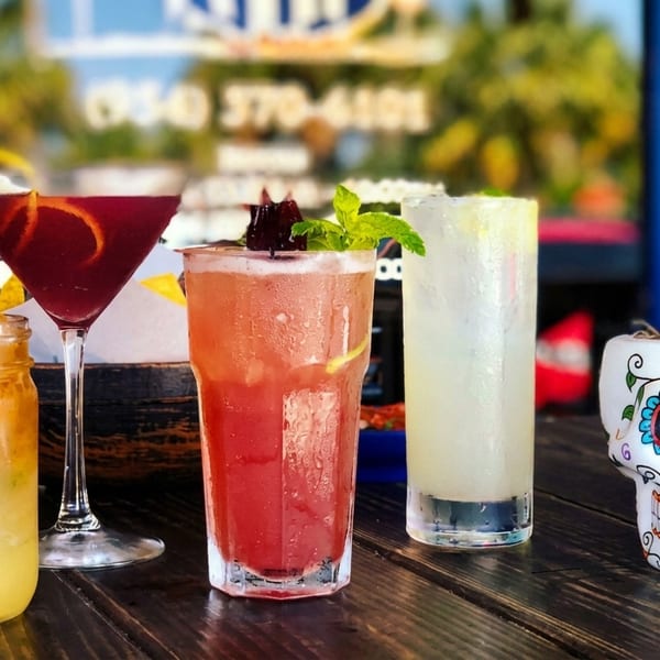 Four colorful cocktails and a decorative sugar skull mug on a wooden table outdoors.