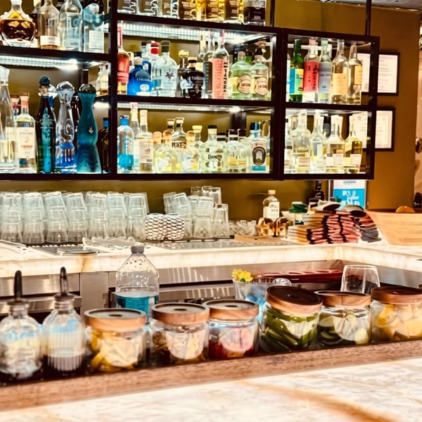 A well-stocked bar with various liquor bottles on shelves, glasses, and jars of garnishes on the counter.