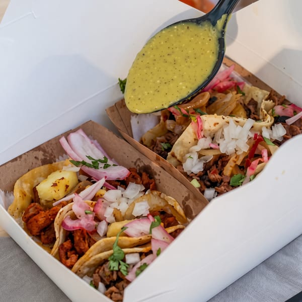 Spoon pouring green sauce over two tacos with meat, onions, and cilantro in a takeout box.