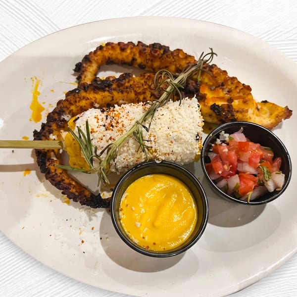 Grilled octopus with elote, pico de gallo, and a yellow dipping sauce on a white plate.