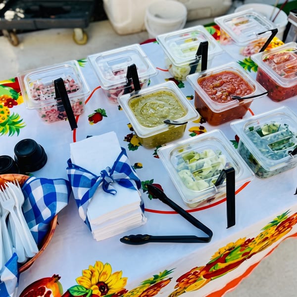 A vibrant tablecloth with fruit and floral patterns holds various salsa and topping containers, with cutlery and napkins.