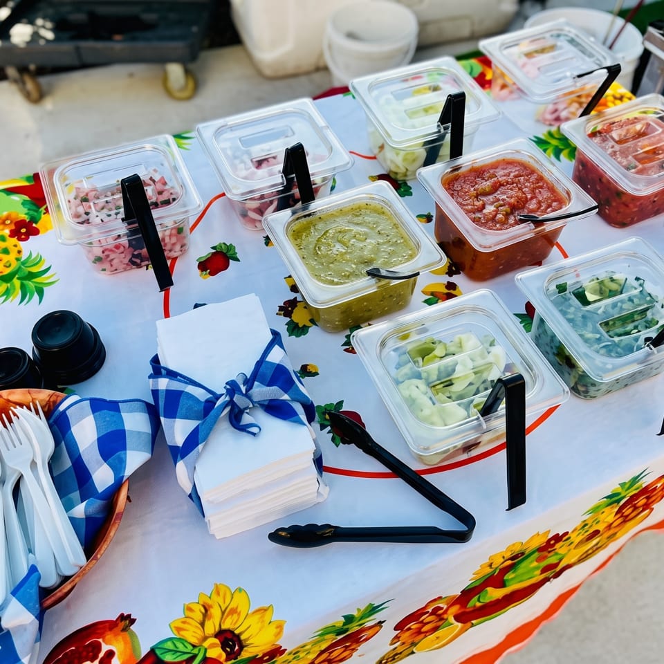A vibrant tablecloth with fruit and floral patterns holds various salsa and topping containers, with cutlery and napkins.