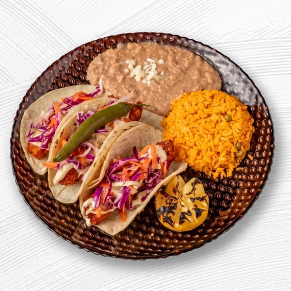 Three fish tacos with cabbage, a grilled pepper, rice, beans, and a charred lemon wedge.