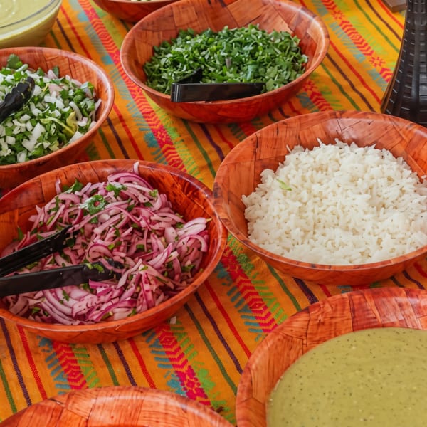 Bowls of rice, chopped onions, cilantro, and green sauces on a colorful striped tablecloth.