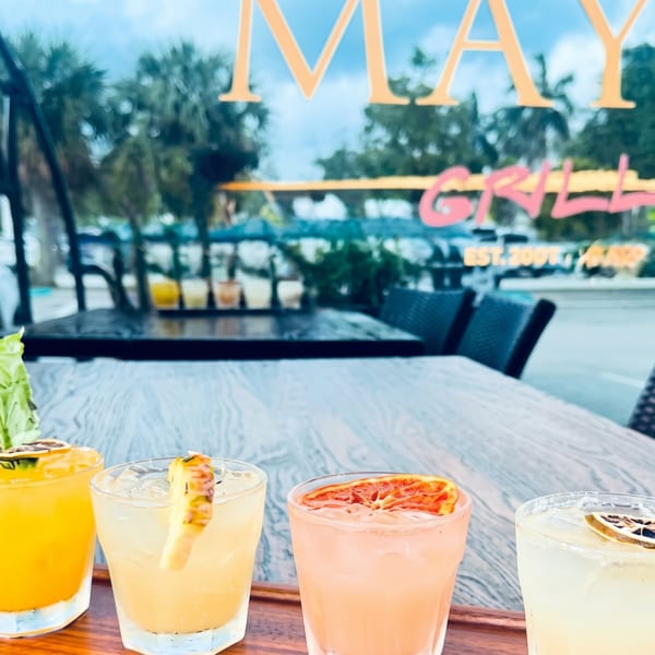 Four colorful cocktails on a wooden tray, with the Maya Grill sign reflected in the window.